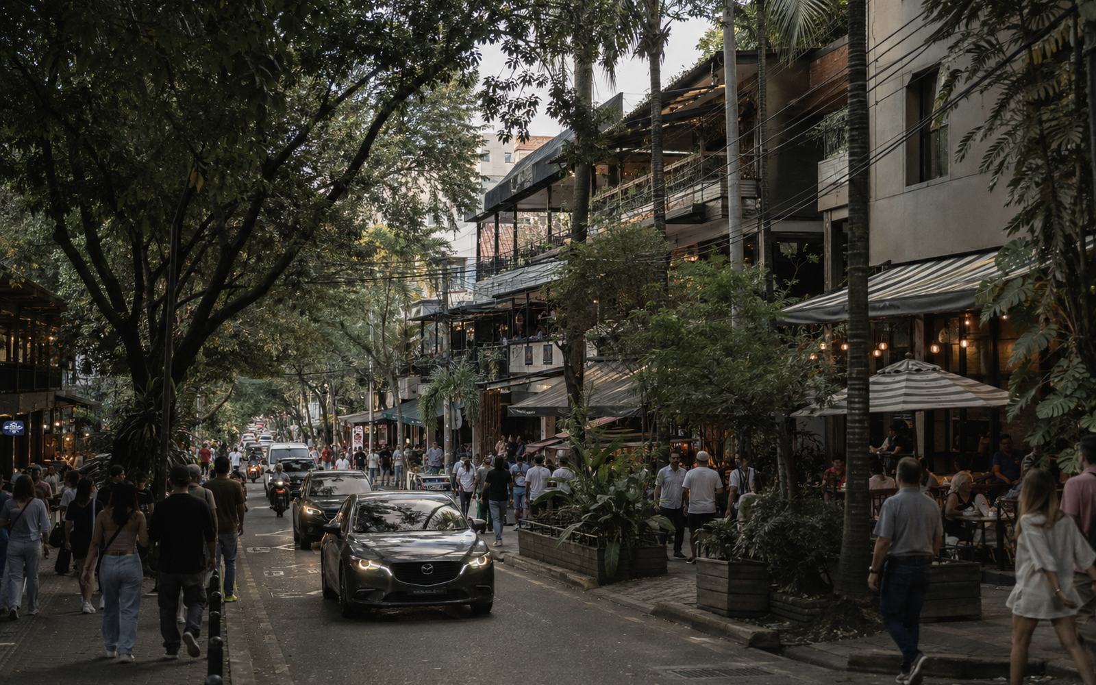 A dense commercial street at dusk with cafés, awnings and heavy foot traffic.